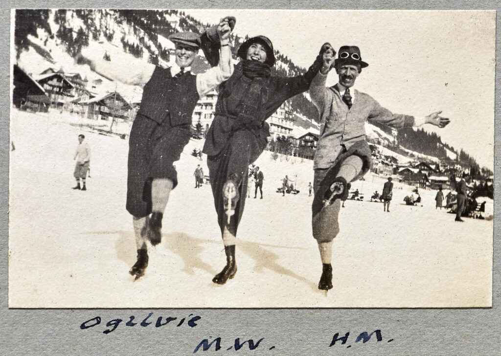 A photograph of three people holding hands wearing ice skates in a snowy Alpine scene